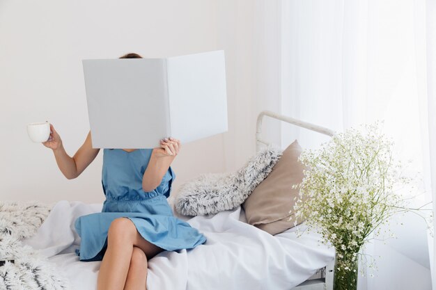 Mujer sentada en la cama cubriendo la cara con el libro tomando café.