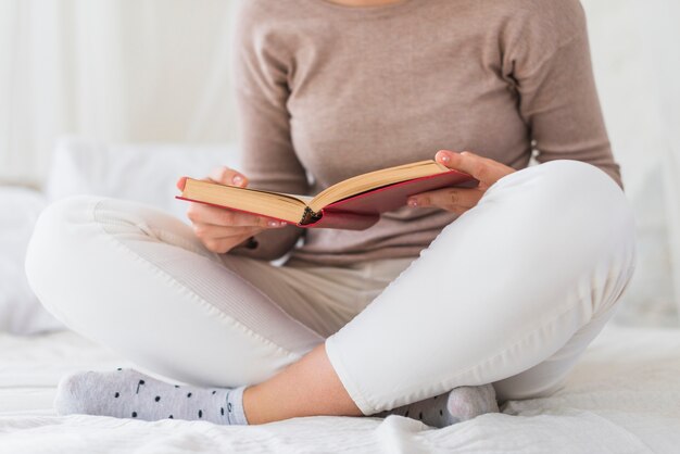 Mujer sentada en la cama con el libro de la celebración de la pierna cruzada