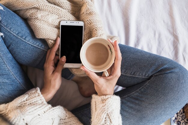 Mujer sentada en la cama con teléfono inteligente y café