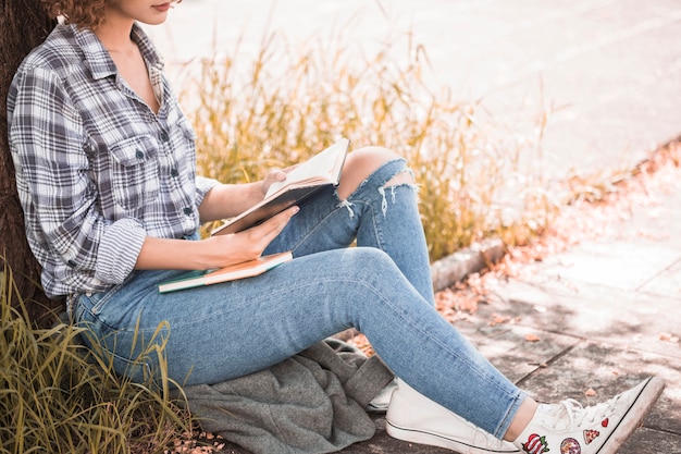 Mujer sentada en el césped cerca de árbol y libro de lectura