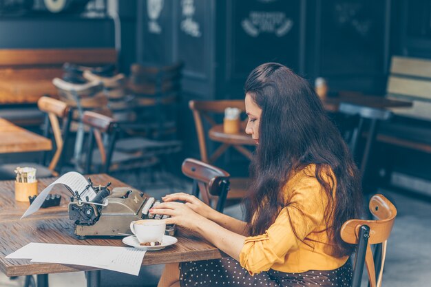 Mujer sentada y escribiendo algo en la máquina de escribir en la terraza del café en la parte superior amarilla y falda larga durante el día y mirando pensativo