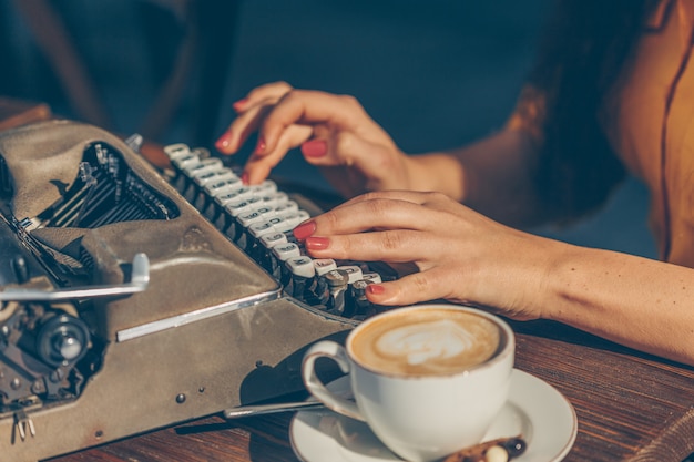Mujer sentada y escribiendo algo en la máquina de escribir en la terraza del café en la parte superior amarilla y falda larga durante el día