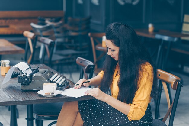 Mujer sentada y escribiendo algo en papel y mirando pensativo y con una falda larga y amarilla en la terraza del café durante el día.