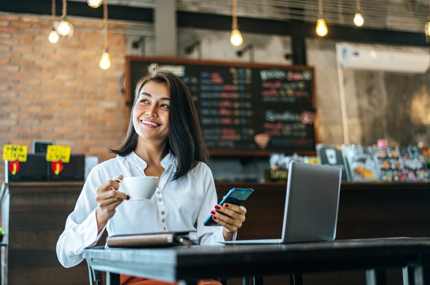 Mujer sentada felizmente trabajando con un teléfono inteligente en una cafetería y portátil.