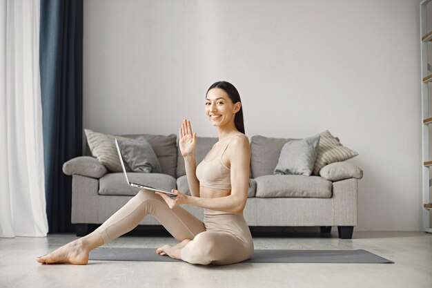 Mujer sentada en un hombre de yoga en leggins y top mientras usa una computadora portátil en casa