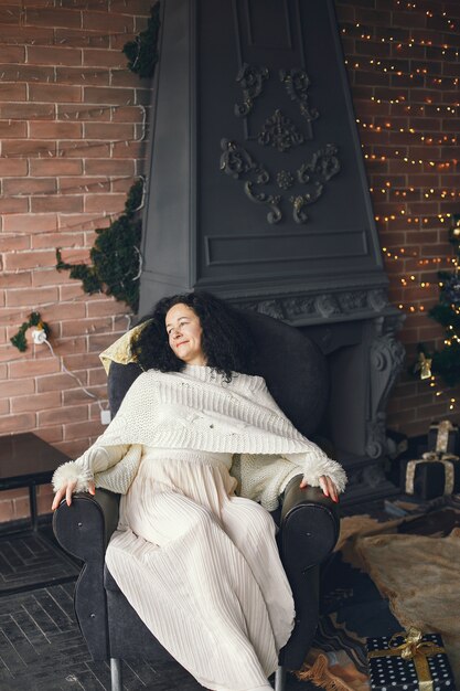 Mujer sentada junto a la chimenea. Dama con un suéter blanco. Morena en un concepto navideño.