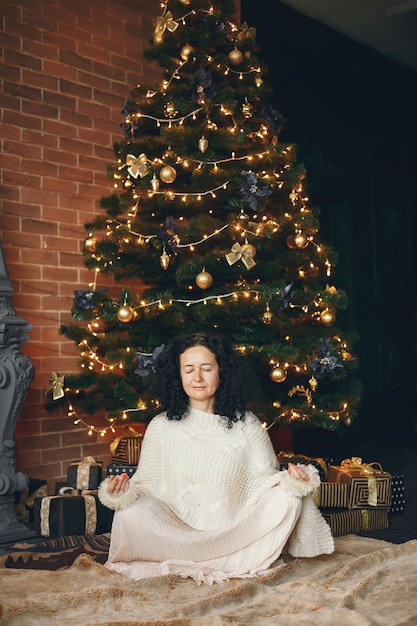 Mujer sentada junto a la chimenea. Dama con un suéter blanco. Morena en un concepto navideño.