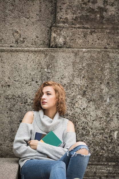 Mujer sentada junto a la pared de piedra y sosteniendo libros