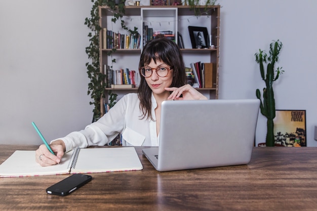 Mujer sentada en mesa con portátil y tomando notas