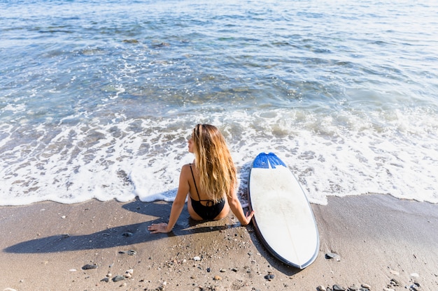 Mujer sentada en la orilla del mar arenoso con tabla de surf