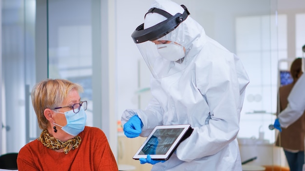 Mujer sentada en una silla en la sala de espera con máscara de protección escuchando médico con aspecto general en tableta en clínica con nueva normalidad. Asistente explicando problema dental durante la pandemia de coronavirus