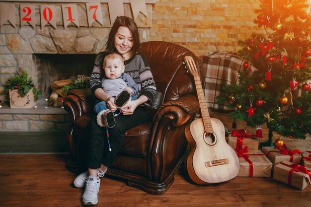 Mujer sentada en un sillón con su bebé y una guitarra, un árbol de navidad y una chimenea de fondo