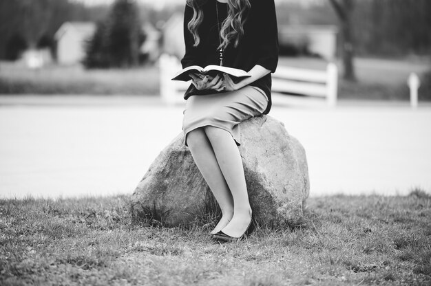 Mujer sentada sobre una roca mientras lee un libro en escala de grises