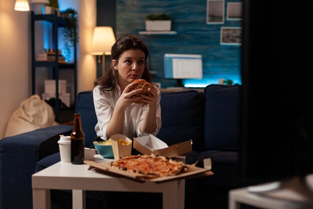 Mujer sentada en el sofá comiendo una deliciosa hamburguesa viendo las noticias en la televisión frente a la mesa de café con pizza grande. Mujer caucásica disfrutando de comida chatarra para llevar.