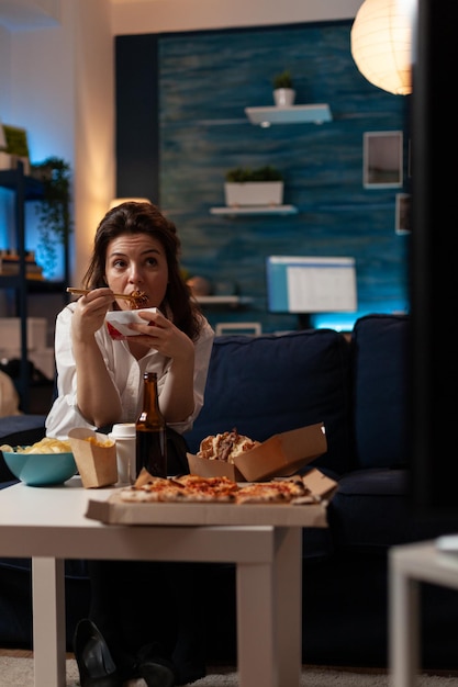 Mujer sentada en el sofá viendo la televisión mientras come fideos asiáticos para llevar con palillos en la sala de estar de casa. Oficinista disfrutando de una deliciosa cena de televisión caja de entrega de comida rápida.