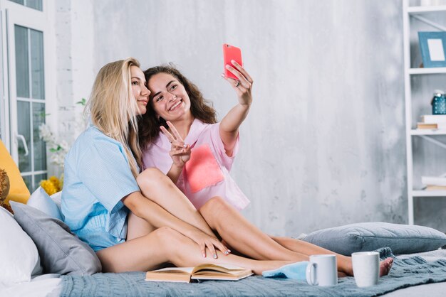 Mujer sentada con su amiga haciendo v signo y tomando selfie en teléfono inteligente