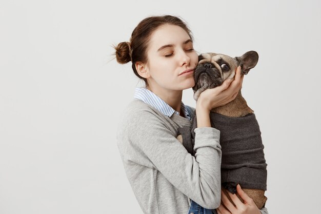 Mujer sentimental presionando su mejilla al bulldog francés que se aisló sobre la pared blanca. Dueño de mascota hembra expresando cuidado y amor abrazando a su perro de pedigrí mientras camina en el parque. Copia espacio