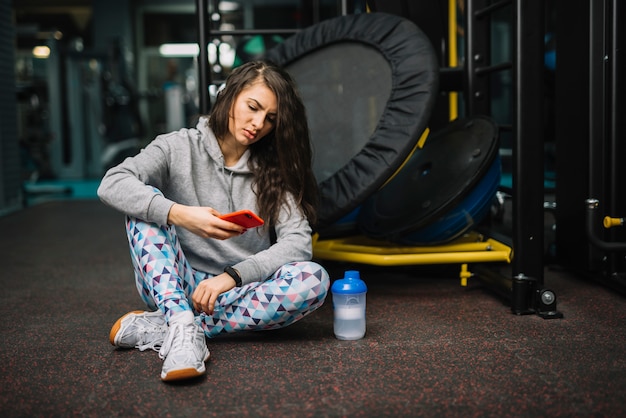 Foto gratuita mujer seria atlética con el teléfono inteligente sentado cerca de la botella en el gimnasio