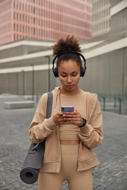 Una mujer seria de cabello rizado vestida con ropa deportiva escucha música a través de auriculares, usa un teléfono inteligente para desplazarse por Internet, lleva karemat enrollado y regresa después de entrenar. Concepto de estilo de vida deportivo.