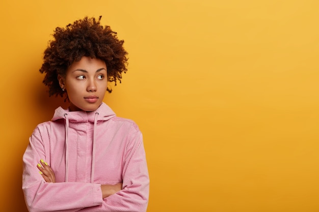 La mujer seria y decidida de pelo rizado se para pensativa, reflexiona sobre algo, mira a un lado, mantiene los brazos cruzados, considera una decisión importante, aislada en la pared amarilla, espacio vacío a un lado.