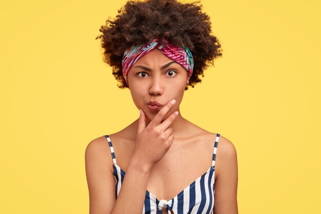 Mujer seria disgustada con peinado rizado afro, sostiene la barbilla, frunce el ceño, vestida con ropa a rayas