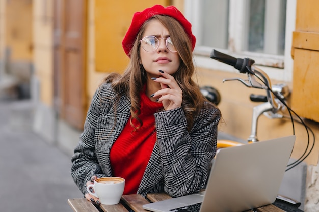 Mujer seria con elegante cabello rizado mirando a la cámara, mientras trabaja con la computadora en la cafetería al aire libre