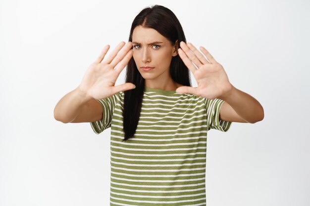 Mujer seria estirar las manos en bloque parando decir no fruncir el ceño y mirando disgustado de pie sobre fondo blanco.