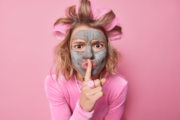 Una mujer seria exige silencio, muestra un signo de silencio que mantiene el dedo índice sobre los labios, usa rulos para hacer rizos, cuenta el secreto de la belleza, se prepara para una cita o una fiesta aislada sobre un fondo rosado.