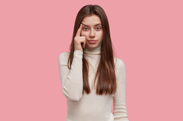 Una mujer seria y guapa se ve con confianza, mantiene el dedo en las sienes, levanta las cejas, tiene el pelo largo y oscuro