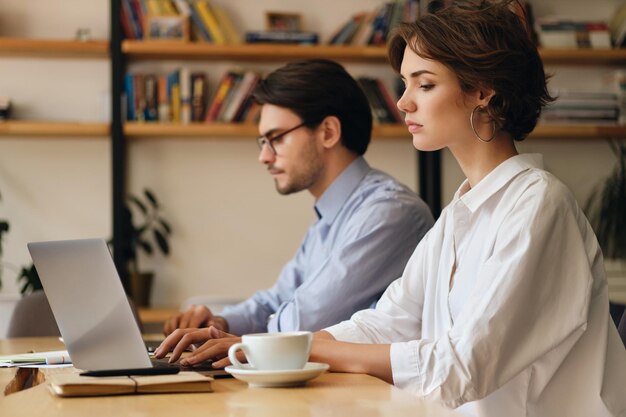 Mujer seria joven sentada en la mesa trabajando cansadamente en una computadora portátil con un colega cerca de la oficina moderna