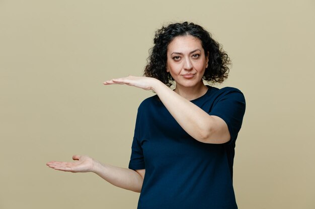 Mujer seria de mediana edad con camiseta mirando a la cámara mostrando un gesto de tamaño aislado en un fondo verde oliva