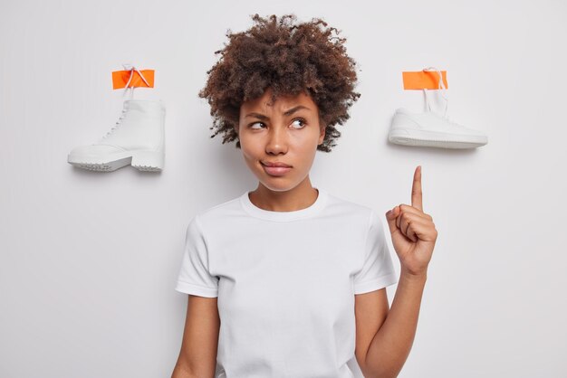 Una mujer seria con el pelo tupido y rizado indica el dedo índice por encima de las poses contra el fondo blanco y anuncia zapatos para usar vestidos de manera informal. Elija entre botas y zapatos deportivos. Toma monocromática.