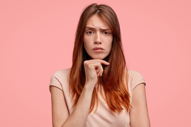 Mujer seria con piel pecosa, mantiene la mano debajo de la barbilla, frunce el ceño, vestida informalmente, aislada sobre fondo rosa.