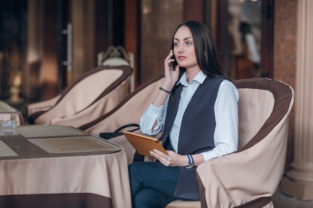 Mujer seria sentada en un restaurante elegante con una tablet y hablando por teléfonomujer seria sentada en un restaurante elegante con una tablet y hablando por teléfono