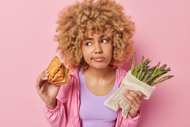 Foto gratuita una mujer seria y vacilante con el pelo rizado elige entre gofres y espárragos se siente confundida vestida con una chaqueta informal enfocada a un lado aislada sobre un fondo rosa dieta o alimentación poco saludable