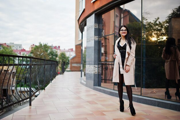 Mujer sexy de cabello negro con gafas y abrigo posado contra el edificio con ventanas modernas