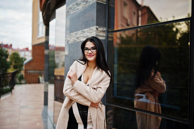 Mujer sexy de cabello negro con gafas y abrigo posado contra el edificio con ventanas modernas