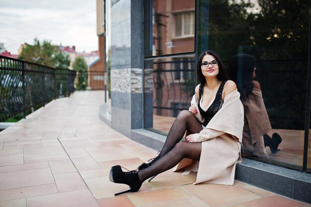 Mujer sexy de cabello negro con gafas y abrigo posado contra el edificio con ventanas modernas