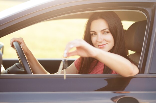 La mujer se sienta en el asiento del conductor, mantiene la mano en el volante, anuncia o vende automóviles. Hermosa mujer morena conduce vehículo