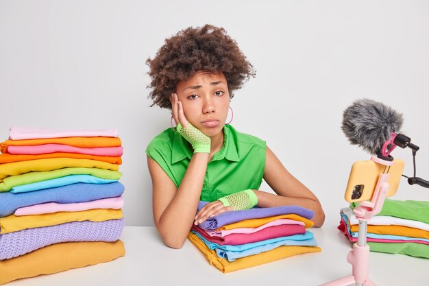 Mujer se siente cansada después de doblar la ropa tiene su propio blog sobre el estilo de vida doméstico se sienta a la mesa frente a la cámara del teléfono inteligente aislada en blanco