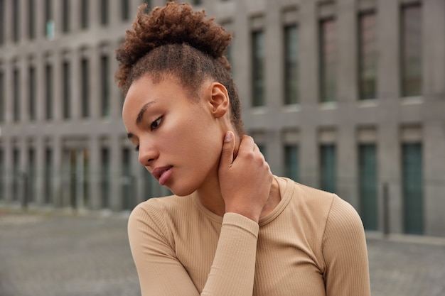 Mujer siente dolor en el cuello después del entrenamiento vestida con ropa deportiva enfocada hacia abajo con poses de expresión seria contra el edificio borroso