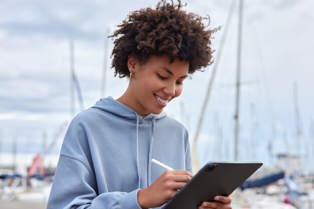 Mujer se siente feliz dibuja bocetos con lápiz óptico en tableta vestida con sudadera con capucha plantea puerto marítimo al aire libre hace dibujos para proyecto futuro