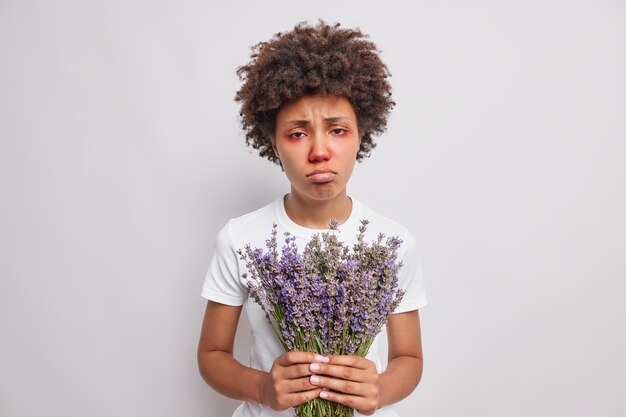 mujer se siente mal sostiene lavanda tiene una reacción alérgica en el polen rojo ojos inflamados picazón nariz que moquea monederos labios usa camiseta casual aislado sobre blanco
