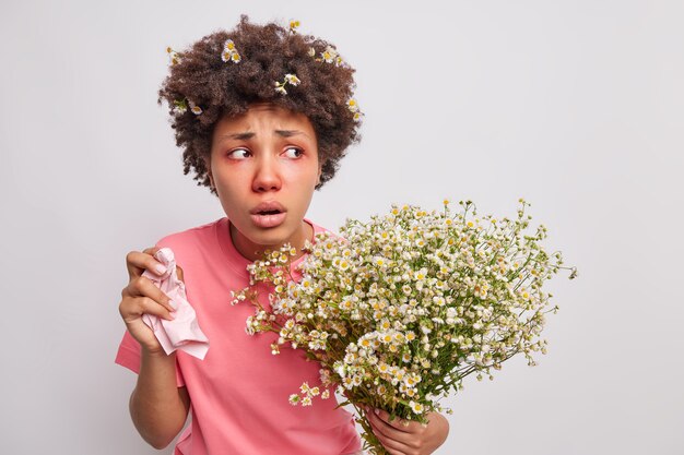 La mujer se siente mal, sufre de síntomas de alergia, sostiene el tejido en la mano, es alérgico a las flores silvestres, sostiene un ramo de poses de manzanilla en blanco