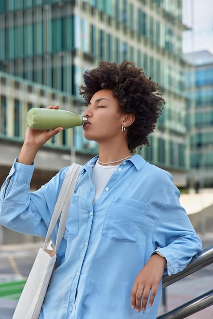 mujer siente sed bebe agua fresca de botella mantiene los ojos cerrados viste camisa casual lleva bolsa refresca con poses de bebidas frías contra edificio moderno exterior