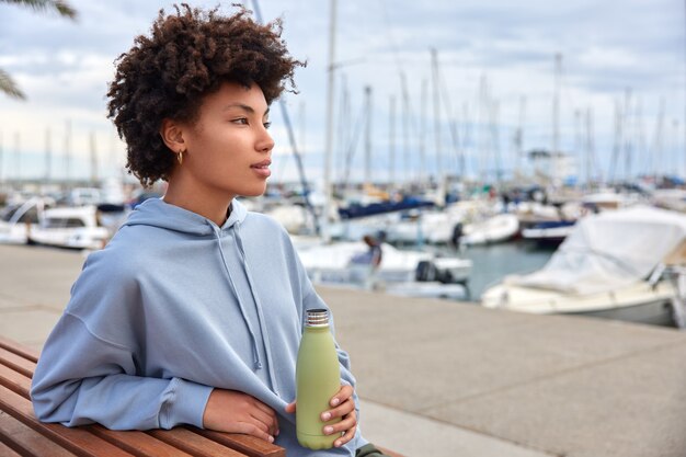 La mujer siente sed sostiene una botella de agua admira la hermosa vista al mar posa en el puerto tiene una expresión pensativa pasa el tiempo libre al aire libre