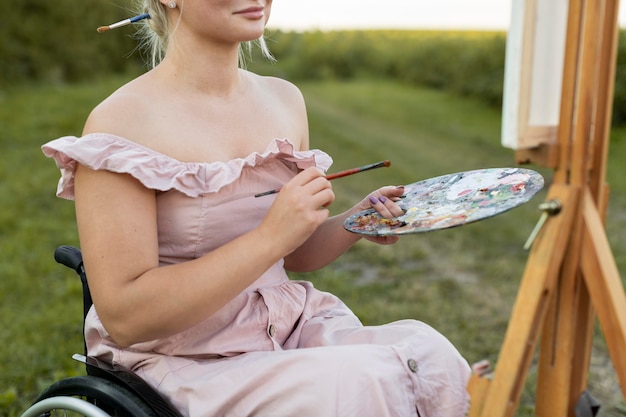 Foto gratuita mujer en silla de ruedas con lienzo y paleta de pintura exterior