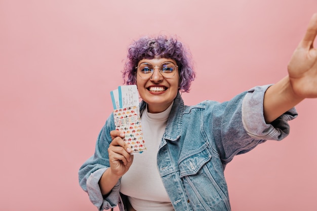 Mujer sinceramente brillante con elegante peinado morado en chaqueta vaquera tiene entradas, sonríe y toma selfie.