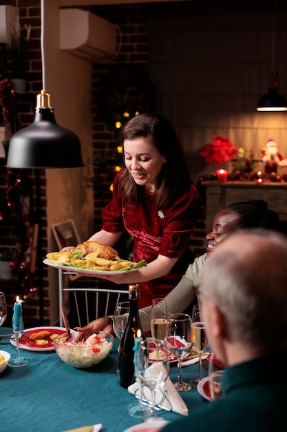 Foto gratuita mujer sirviendo comida festiva en la mesa, celebrando la cena de navidad con diversos amigos y familiares en casa. la gente se siente feliz comiendo comida casera y bebiendo vino durante el evento de víspera de navidad.