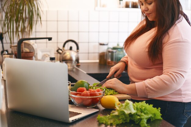 Mujer con sobrepeso que usa la computadora portátil para ver la receta en video mientras hace una ensalada de aguacate con vitamina vegana, cortando lechuga en una tabla de cortar de madera. Concepto de comida sana, pérdida de peso, dieta y nutrición